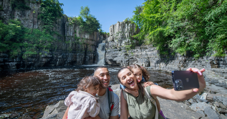 Family taking a selfie at High Force Waterfall on a bright sunny day.